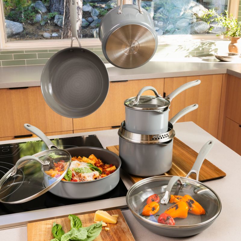 Gray Circulon cookware set in a sunny kitchen: pasta in a sauté pan on an induction cooktop, stacked saucepans, hanging pans and peppers in a lidded frying pan