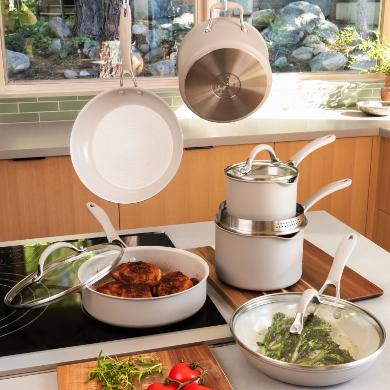 Taupe cookware in a kitchen: frying pan and saucepan hanging, stacked saucepans, chicken in a sauté pan on the cooktop, broccolini in a covered frying pan