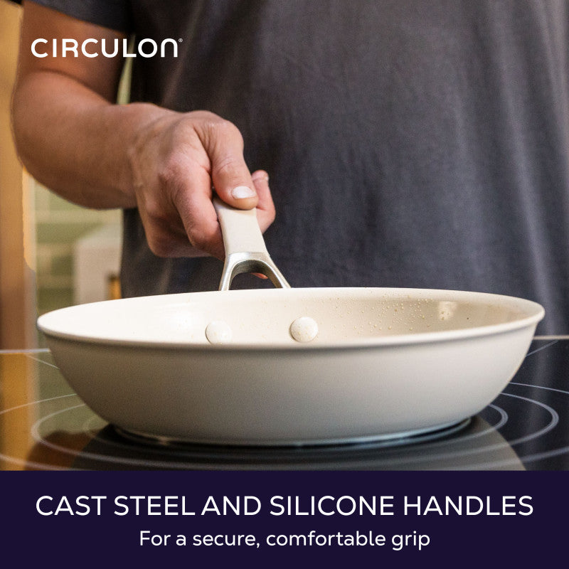 Cast steel and silicone handles for a secure, comfortable grip: hand holding a taupe frying pan by its handle over a cooktop