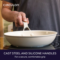 Cast steel and silicone handles for a secure, comfortable grip: hand holding a taupe frying pan by its handle over a cooktop