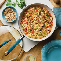 Overhead view of the marine blue stir fry pan of shrimp pasta with peppers, glass lid and a bowl of slivered almonds beside it