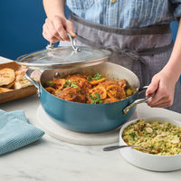 Person lifting the glass lid off an agave blue casserole of braised chicken with olives and lemon, beside couscous and flatbread