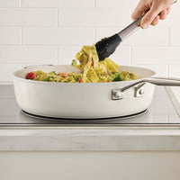 Porcelain white sauté pan of pesto pappardelle with tomatoes on an induction cooktop, lifted with tongs