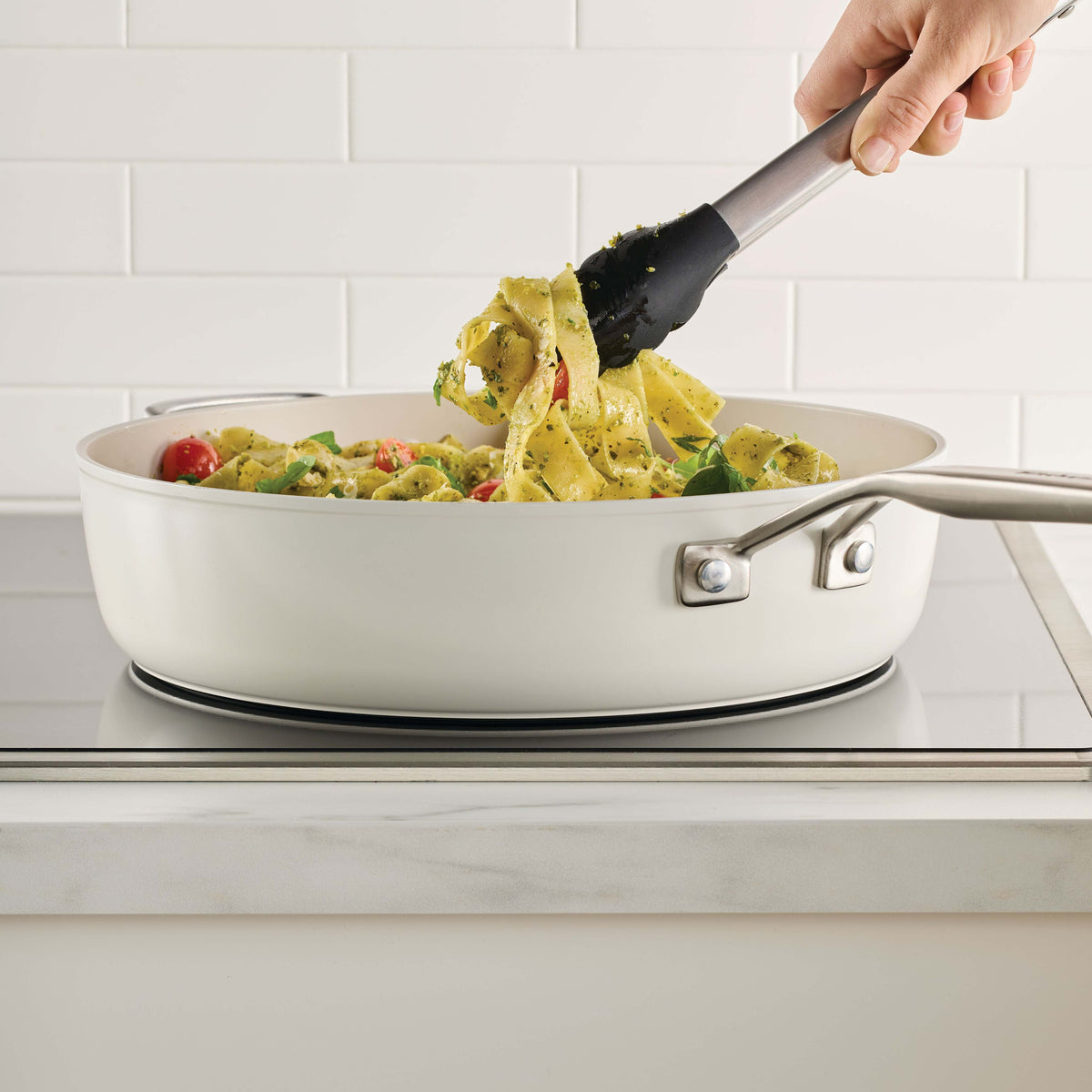 Porcelain white sauté pan of pesto pappardelle with tomatoes on an induction cooktop, lifted with tongs