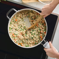 Porcelain white sauté pan of risotto with peas and carrots on an induction cooktop, stirred with a wooden spoon
