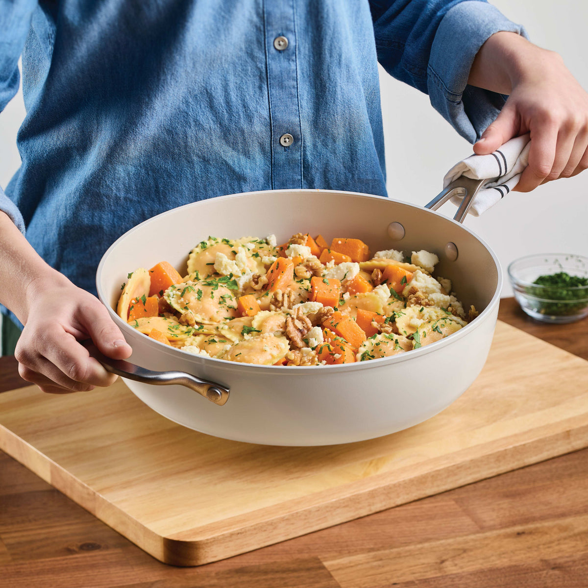 Porcelain white sauté pan of ravioli with butternut squash, walnuts and feta, held by its long handle and helper handle over a wooden cutting board