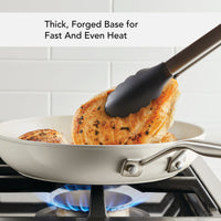 Thick, forged base for fast and even heat: tongs turning chicken breasts in the porcelain white ceramic frying pan over a gas flame