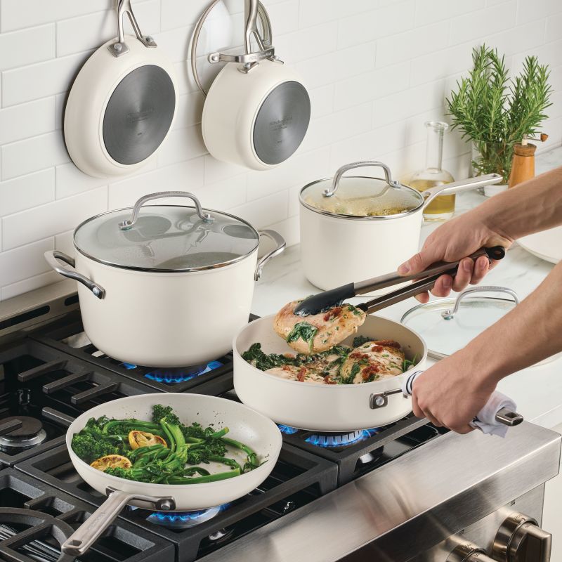 Porcelain white cookware on a gas range: tongs turning chicken in a sauté pan beside broccolini in a frying pan, with two pans hanging on the tile wall