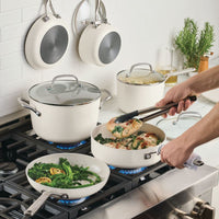 Porcelain white cookware on a gas range: tongs turning chicken in a sauté pan beside broccolini in a frying pan, with two pans hanging on the tile wall