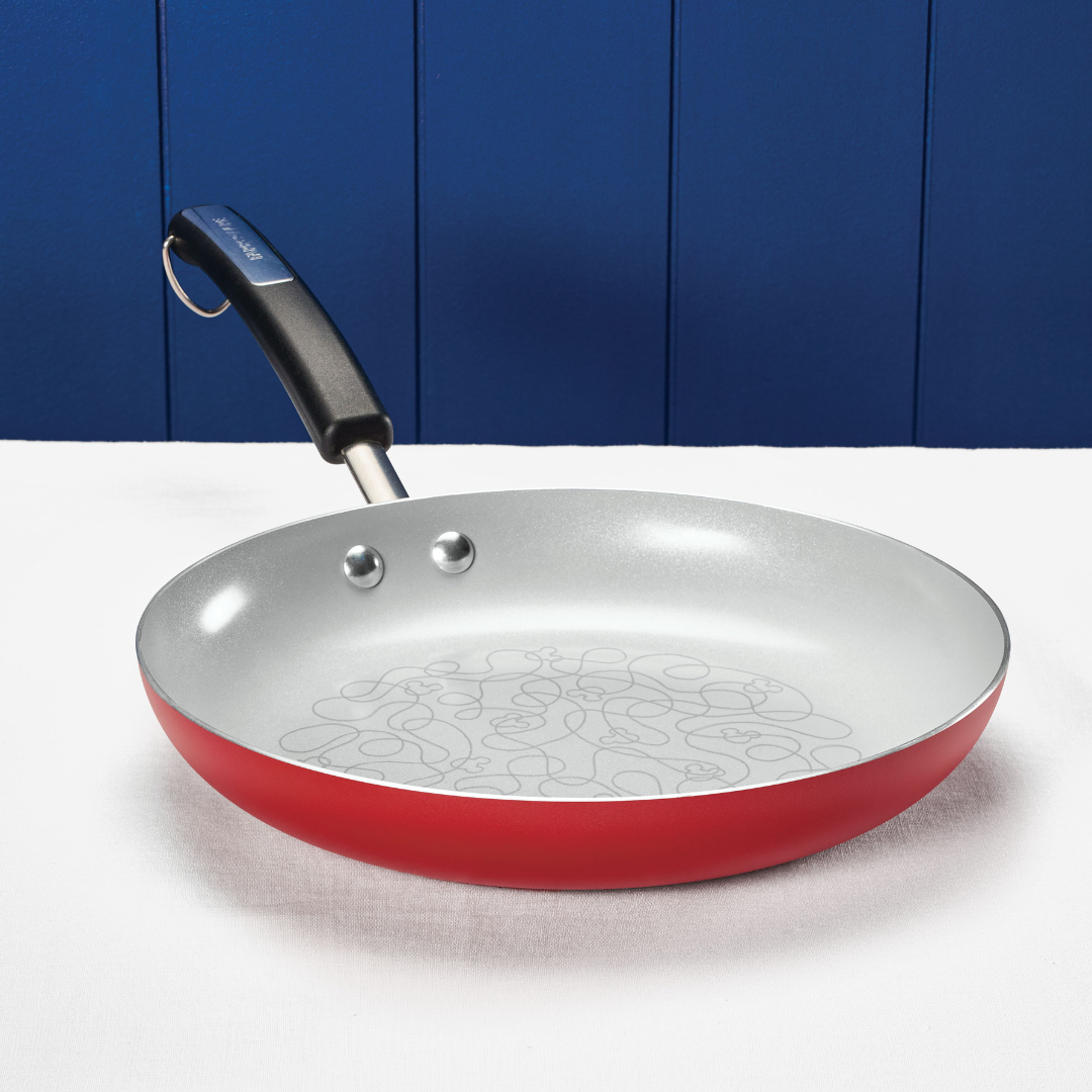Red ceramic nonstick frying pan with a Mickey Mouse doodle pattern inside, on a white tablecloth
