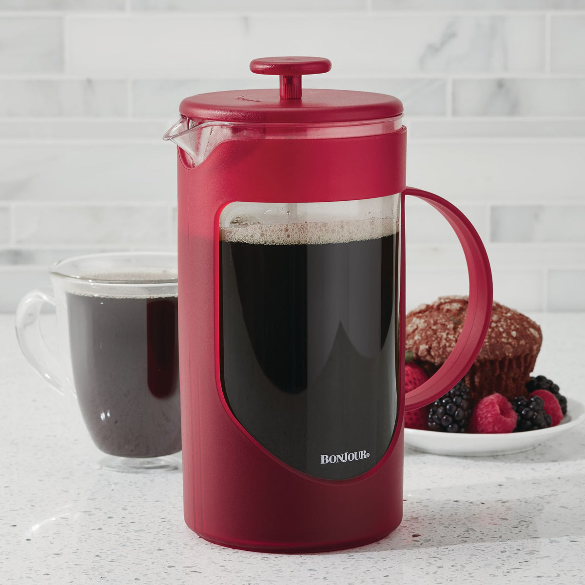 Red French press of coffee on a counter with a glass mug of coffee and a muffin with berries